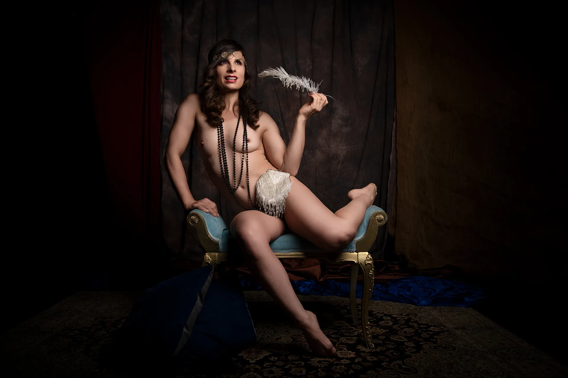 Nude female model seated on a stool in a theatrical boudoir setting, posed with elegance and confidence under dramatic studio lighting.