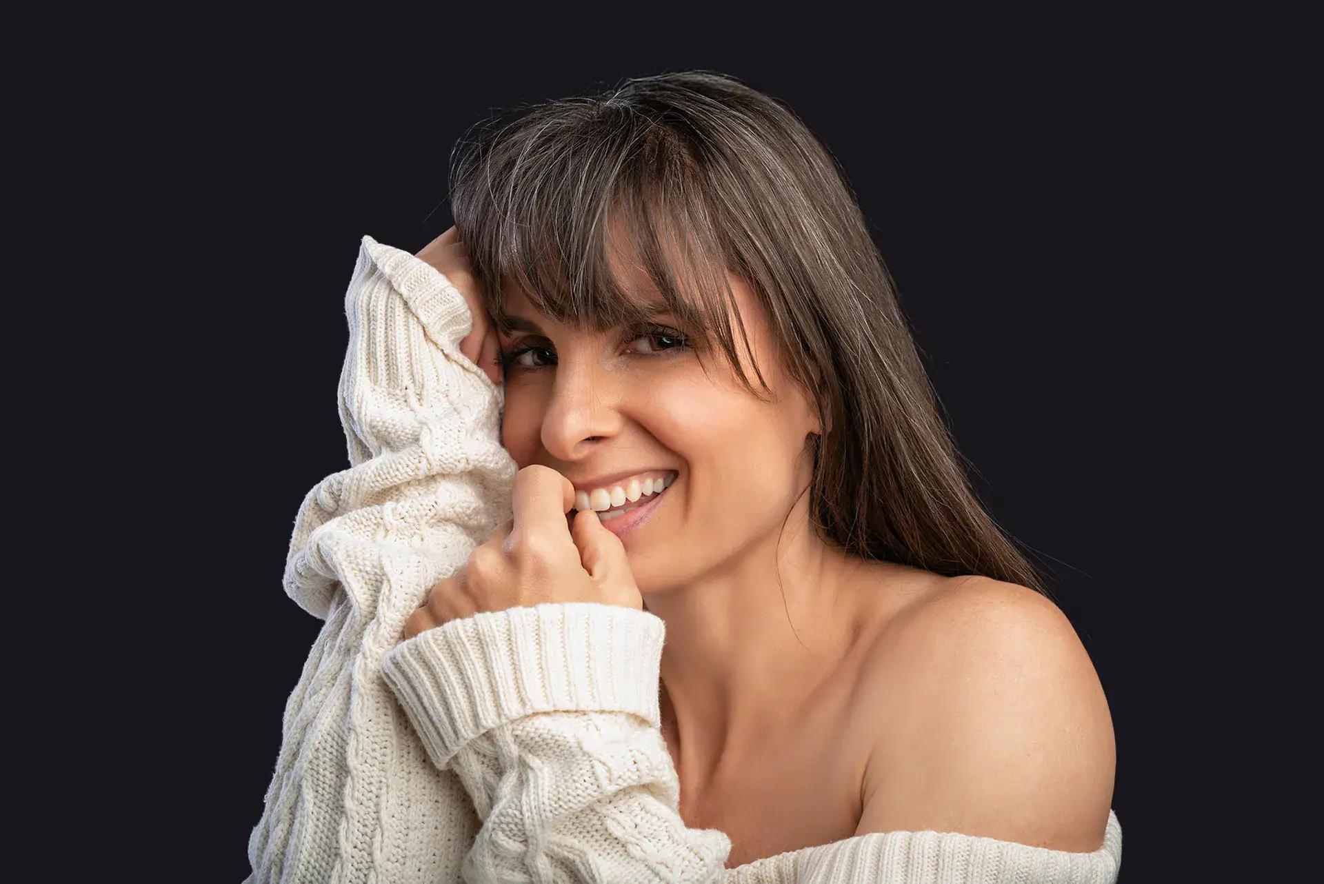 Relaxed boudoir portrait of smiling woman in off-shoulder knitwear Smiling woman wearing white knit sweater against a black backdrop