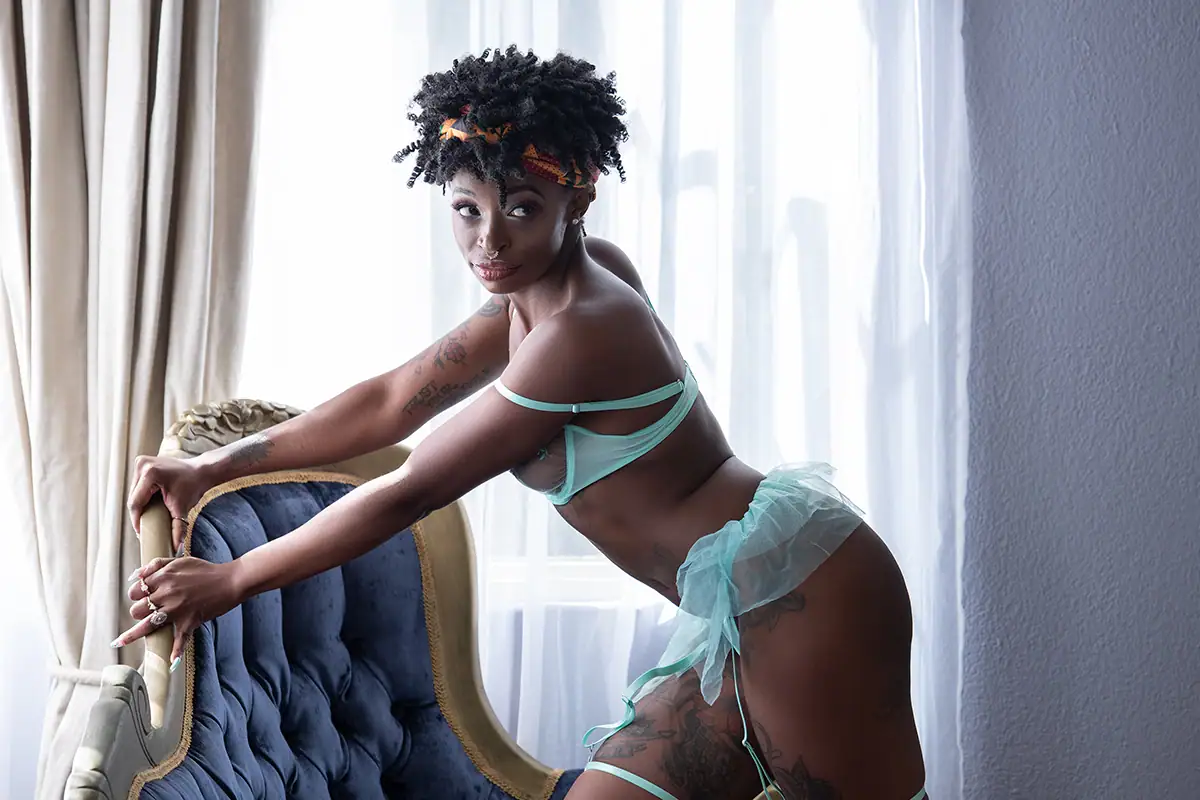 Woman in a revealing bikini kneeling in front of a washing machine, peeking inside with a playful pose, set in a laundry room with a blue wall and concrete floor