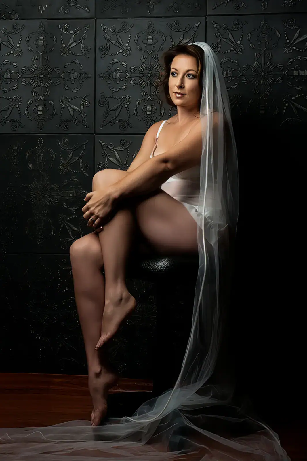 Bridal Boudoir Beneath the Veil Artistic bridal nude portrait of a seated woman partially draped in her wedding veil against a dark studio backdrop