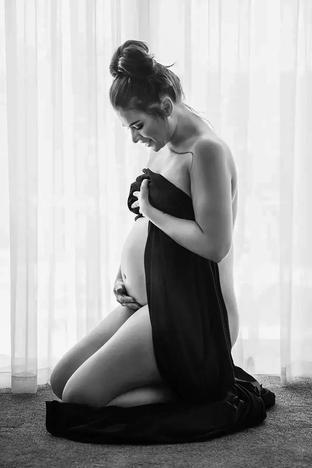 Black and White Maternity Boudoir Portrait Pregnant woman kneeling beside a bright window in an elegant black and white maternity portrait