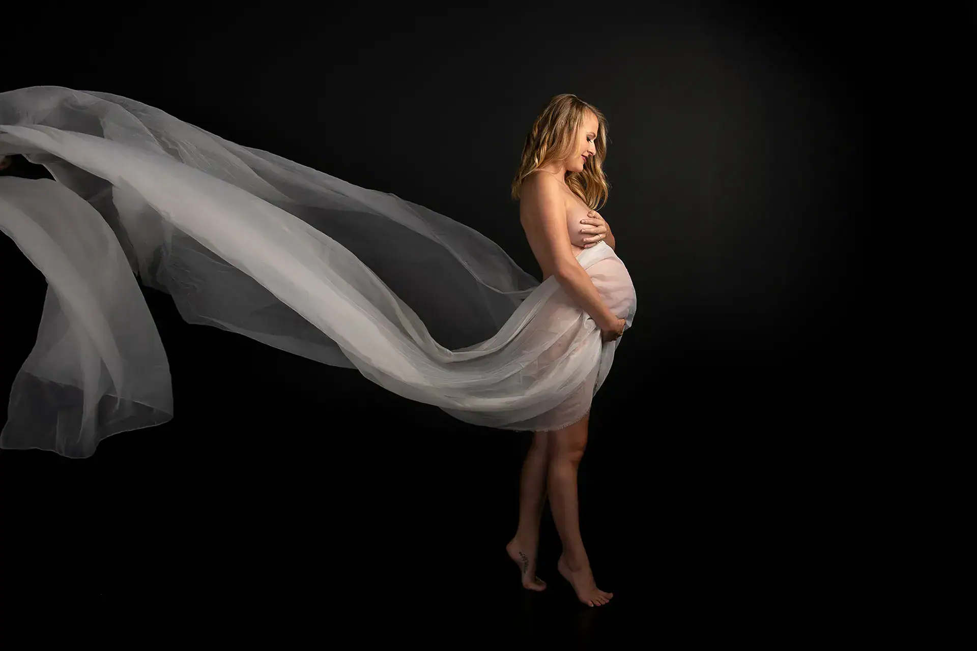 Dramatic Flowing Fabric Maternity Portrait Pregnant woman with sweeping white fabric photographed against a dark studio background